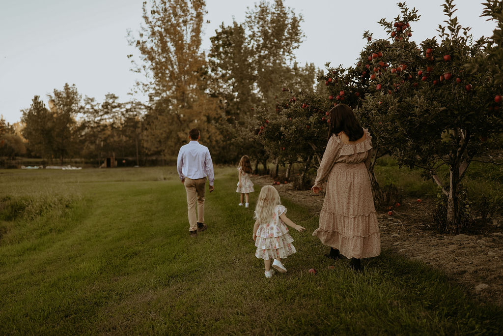 A family walks through an orchard, with the mother and youngest child near apple trees and the father and older child ahead on a grassy path.