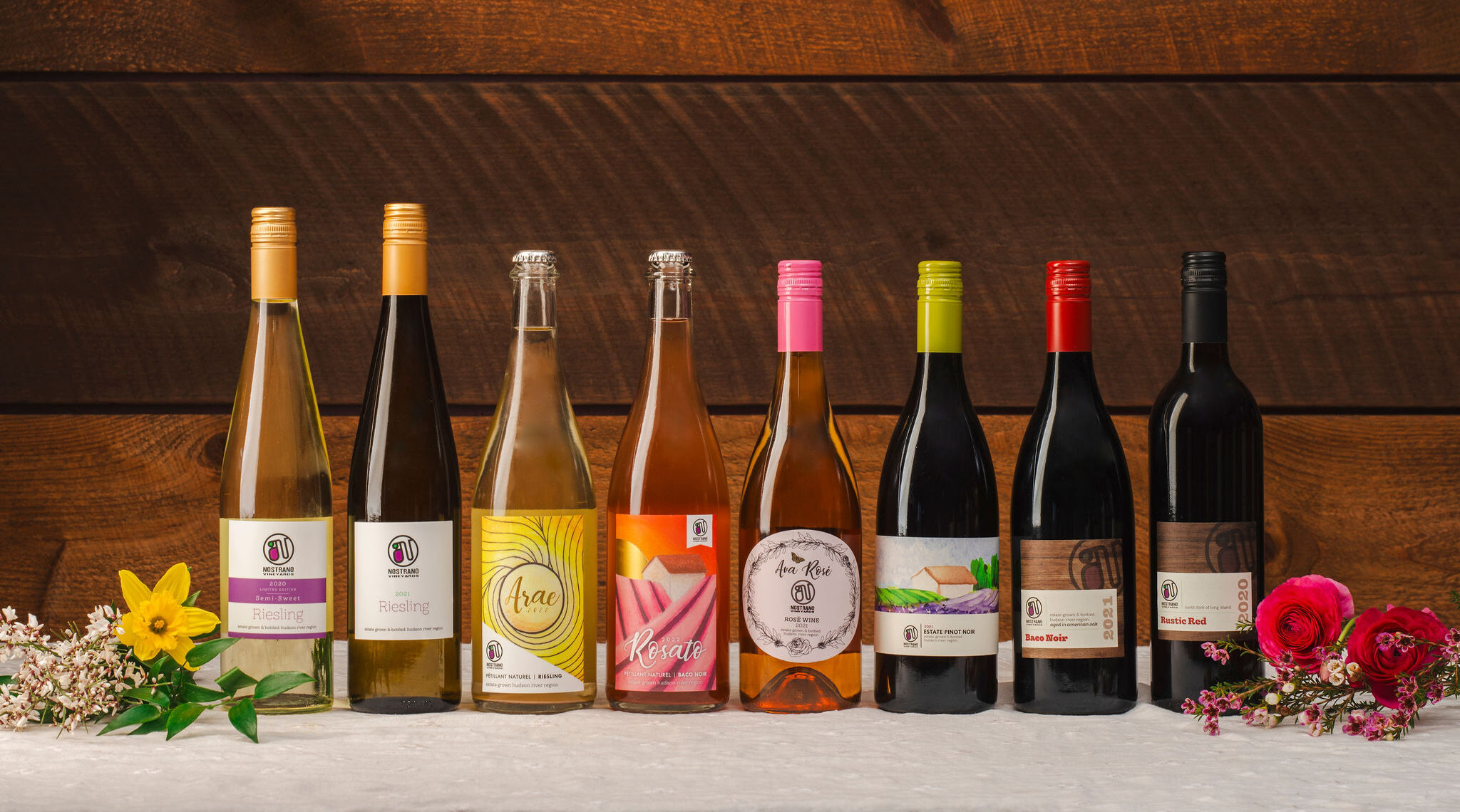 A variety of eight wine bottles displayed in a row on a table with flowers, against a wooden background.