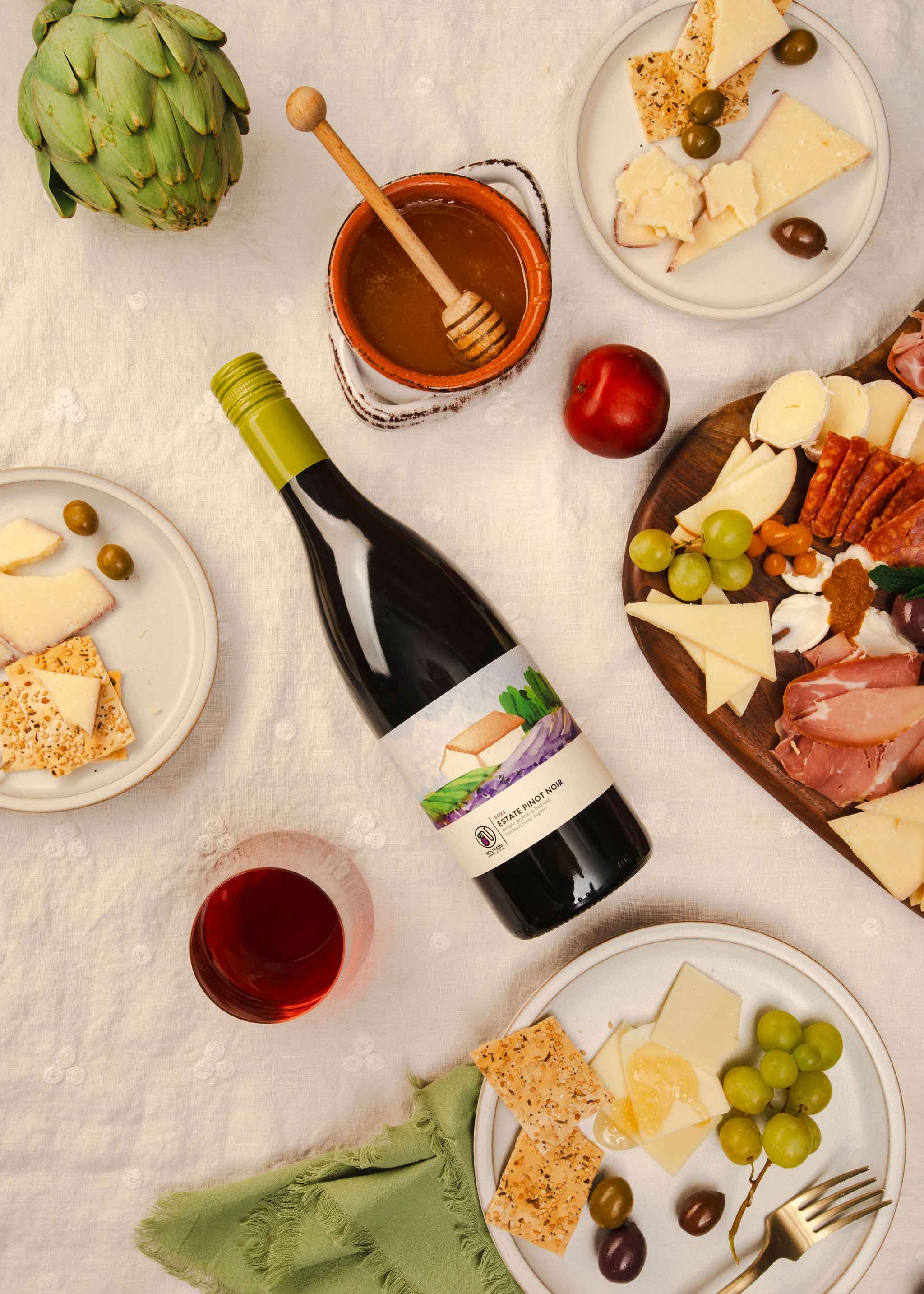 A bottle of Pinot Noir wine with cheese, grapes, olives, crackers, charcuterie, and honey, arranged on a table for a picnic.