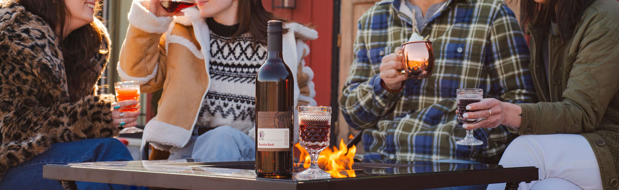 Friends gathered around a fire pit, laughing and drinking wine and cocktails on a sunny day outside a red cabin.