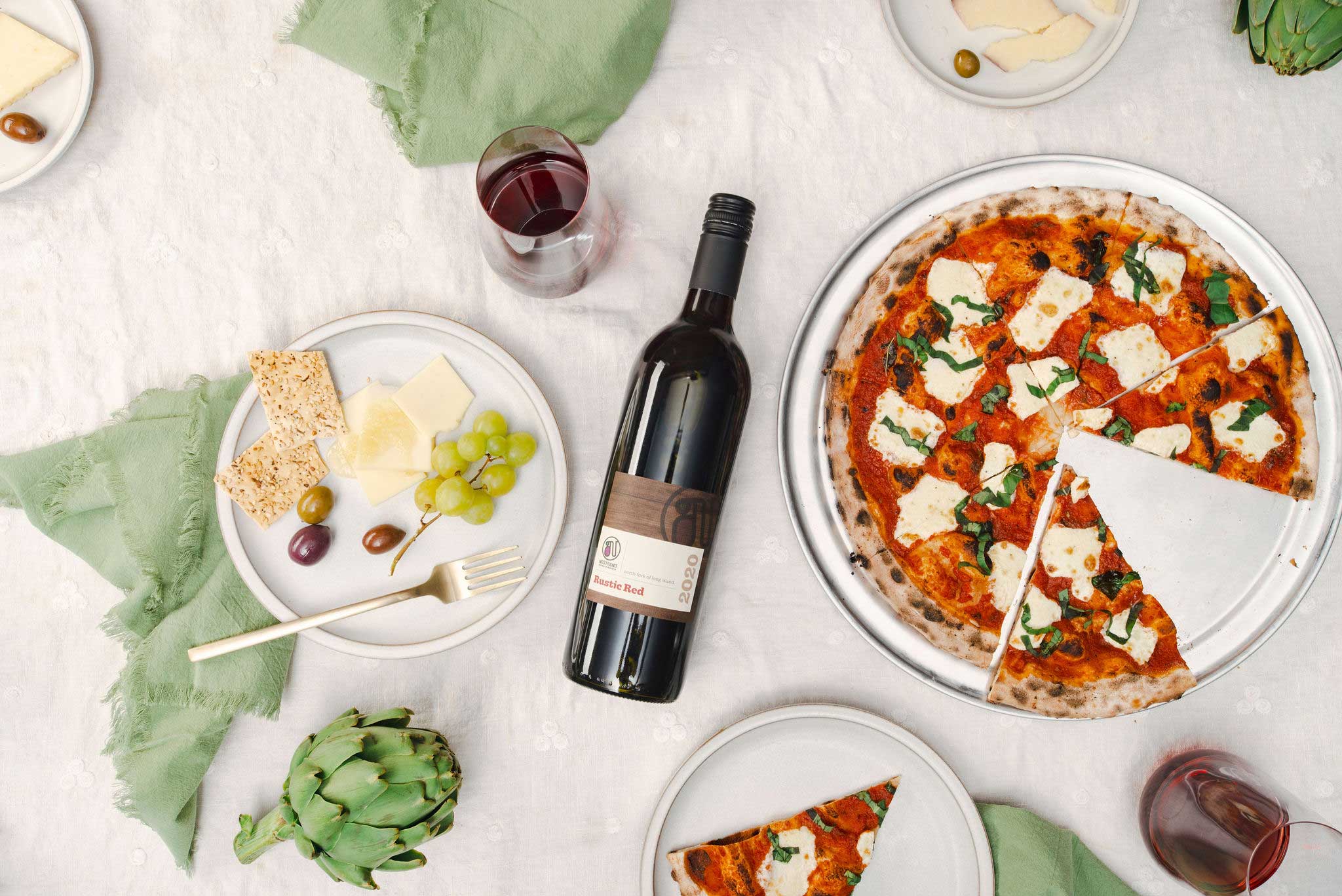 A table set with a margherita pizza, a bottle of red wine, cheese, crackers, olives, and grapes on plates.