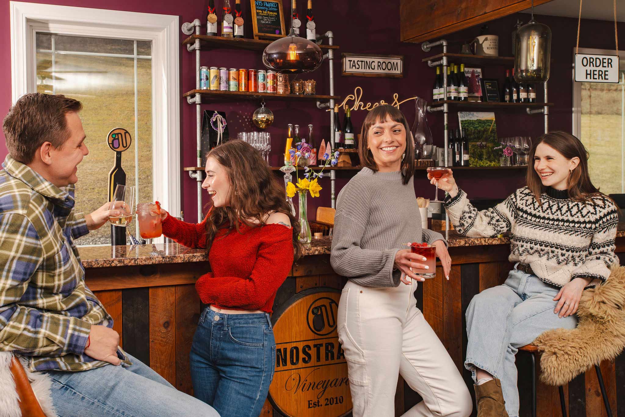 Group of friends laughing and toasting with drinks inside a cozy tasting room at a vineyard.