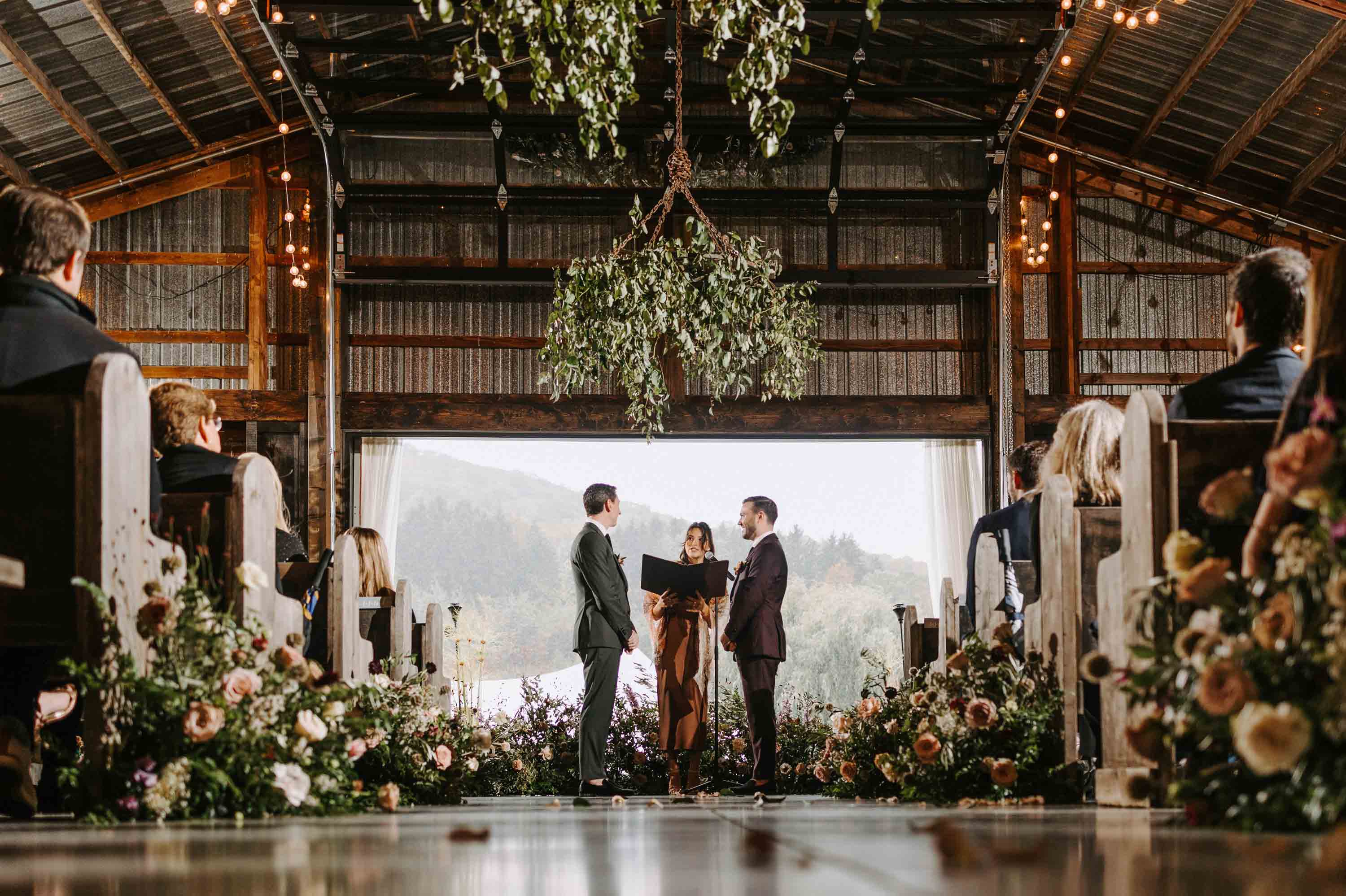 Two grooms stand at the altar during an indoor wedding ceremony, surrounded by flowers and seated guests in a rustic barn.